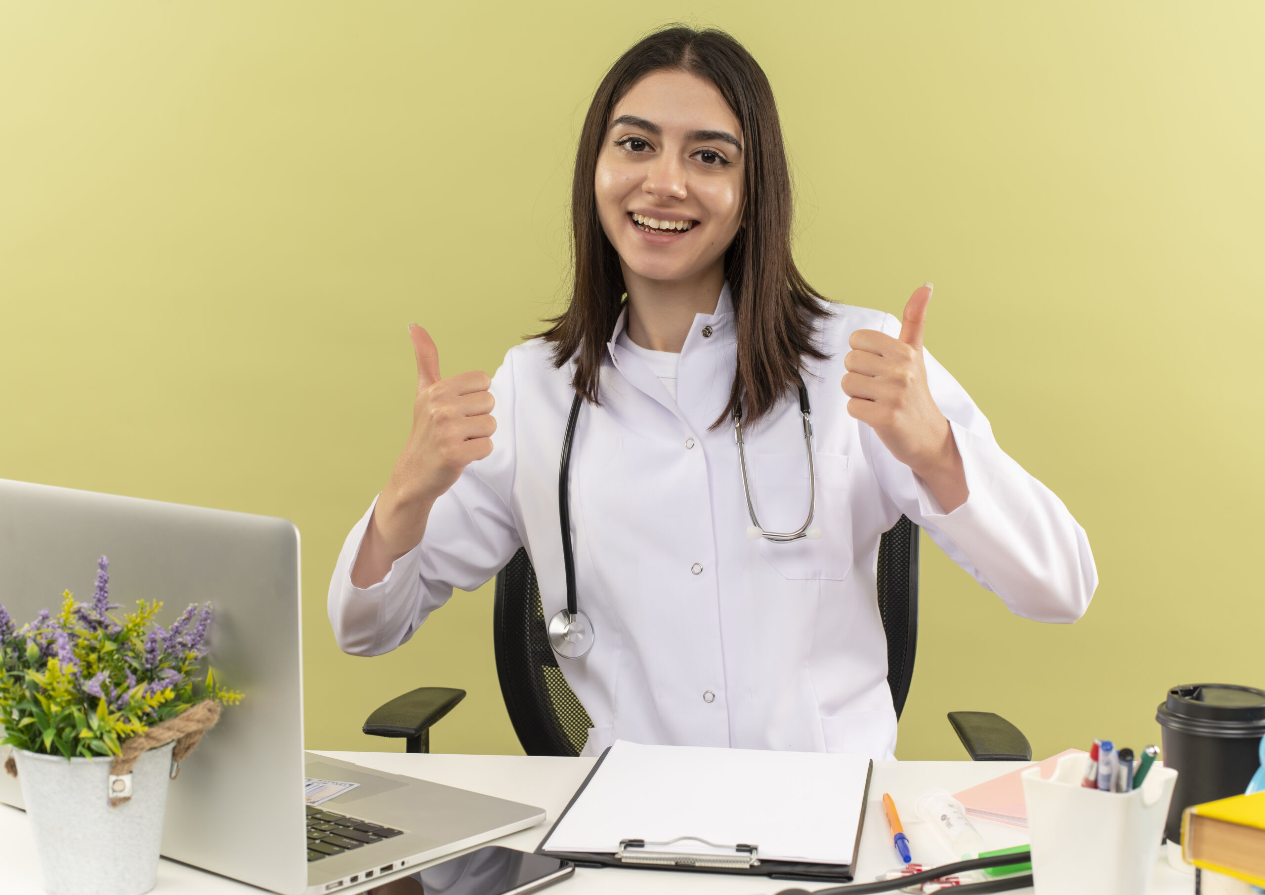 young-female-Primary-care-doctor-white-coat-with-stethoscope-around-her-neck-looking-front-smiling-cheerfully-showing-thumbs-up-sitting-table-with-laptop-light-wall